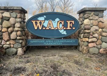 A large, oval-shaped entrance sign for the Wawasee Area Conservancy Foundation (WACF), mounted between two rustic stone pillars. The sign is dark teal with "W.A.C.F." written in bold gold letters over a light blue map of Lake Wawasee. Below the oval, a rectangular plaque displays the full name of the organization. The sign is surrounded by dry, dormant grass and tall reeds under a cloudy sky.