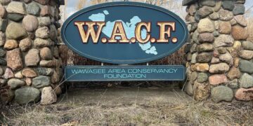 A large, oval-shaped entrance sign for the Wawasee Area Conservancy Foundation (WACF), mounted between two rustic stone pillars. The sign is dark teal with "W.A.C.F." written in bold gold letters over a light blue map of Lake Wawasee. Below the oval, a rectangular plaque displays the full name of the organization. The sign is surrounded by dry, dormant grass and tall reeds under a cloudy sky.