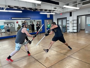 In a spacious, mirrored gym with blue walls and a polished wooden floor, two men are mid-action in a lightsaber duel. On the left, a man in a grey tank top and patterned shorts lunges forward, gripping a lightsaber with a white blade and red detailing. On the right, a man in a black T-shirt and shorts lunges back, holding a solid white lightsaber. The room is brightly lit by overhead fluorescent lights, and several other people can be seen in the background and reflected in the large mirrors, some observing the match.