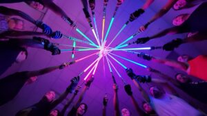 Low-angle, POV shot from the center of a circle of diverse people, all pointing brightly lit lightsabers of various colors (blue, green, purple, and red) toward a single central point in the ceiling. The scene is bathed in a vibrant, neon purple and pink glow, and many of the participants are wearing athletic gear and gloves.