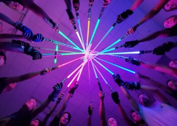 Low-angle, POV shot from the center of a circle of diverse people, all pointing brightly lit lightsabers of various colors (blue, green, purple, and red) toward a single central point in the ceiling. The scene is bathed in a vibrant, neon purple and pink glow, and many of the participants are wearing athletic gear and gloves.