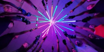 Low-angle, POV shot from the center of a circle of diverse people, all pointing brightly lit lightsabers of various colors (blue, green, purple, and red) toward a single central point in the ceiling. The scene is bathed in a vibrant, neon purple and pink glow, and many of the participants are wearing athletic gear and gloves.