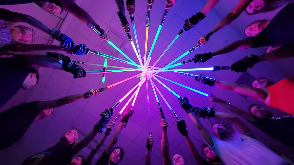 Low-angle, POV shot from the center of a circle of diverse people, all pointing brightly lit lightsabers of various colors (blue, green, purple, and red) toward a single central point in the ceiling. The scene is bathed in a vibrant, neon purple and pink glow, and many of the participants are wearing athletic gear and gloves.