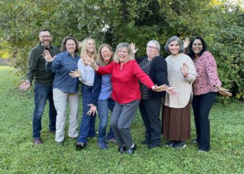 Eight people stand together in a grassy area in front of lush green foliage, smiling and waving at the camera