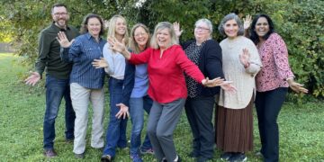 Eight people stand together in a grassy area in front of lush green foliage, smiling and waving at the camera