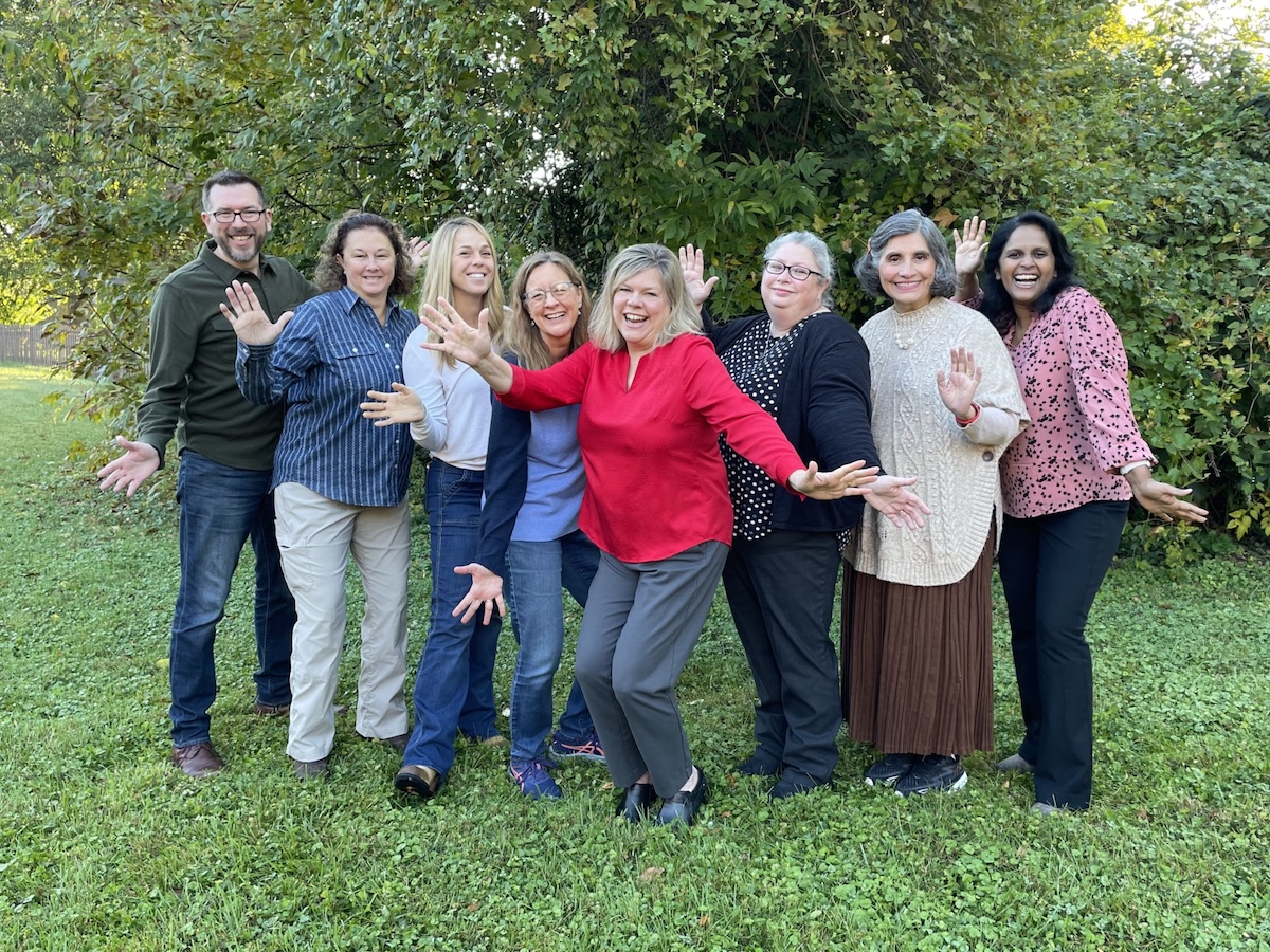 Eight people stand together in a grassy area in front of lush green foliage, smiling and waving at the camera