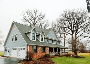 A large, two-story farmhouse with light blue horizontal siding and a red brick facade on the lower right level sits under an overcast sky. The house features a prominent grey shingle roof with three dormer windows and a spacious wraparound front porch adorned with festive holiday garland. Two white garage doors are situated on the left side of the home. Bare, tall trees frame the property, which is surrounded by a green lawn and a glimpse of a nearby field.