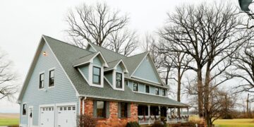 A large, two-story farmhouse with light blue horizontal siding and a red brick facade on the lower right level sits under an overcast sky. The house features a prominent grey shingle roof with three dormer windows and a spacious wraparound front porch adorned with festive holiday garland. Two white garage doors are situated on the left side of the home. Bare, tall trees frame the property, which is surrounded by a green lawn and a glimpse of a nearby field.