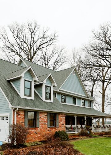 A large, two-story farmhouse with light blue horizontal siding and a red brick facade on the lower right level sits under an overcast sky. The house features a prominent grey shingle roof with three dormer windows and a spacious wraparound front porch adorned with festive holiday garland. Two white garage doors are situated on the left side of the home. Bare, tall trees frame the property, which is surrounded by a green lawn and a glimpse of a nearby field.