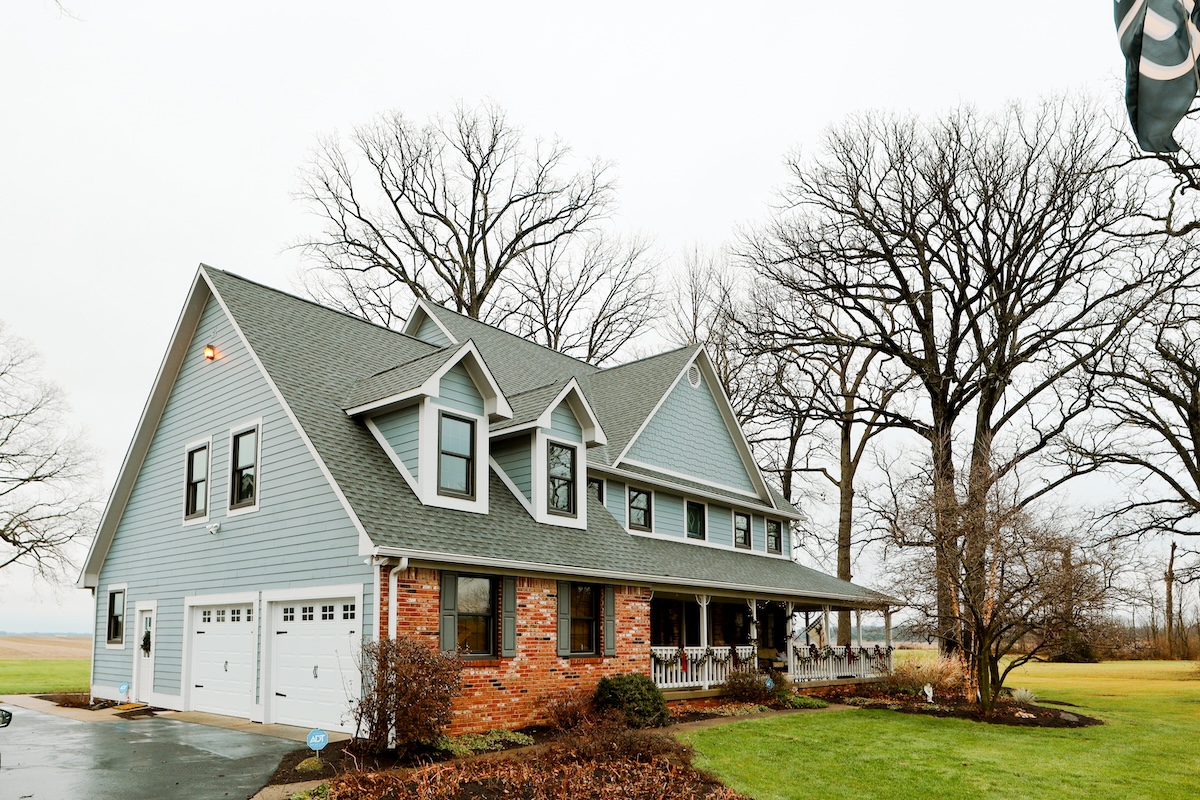 A large, two-story farmhouse with light blue horizontal siding and a red brick facade on the lower right level sits under an overcast sky. The house features a prominent grey shingle roof with three dormer windows and a spacious wraparound front porch adorned with festive holiday garland. Two white garage doors are situated on the left side of the home. Bare, tall trees frame the property, which is surrounded by a green lawn and a glimpse of a nearby field.