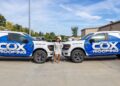 A woman in a light-colored dress and heels stands confidently between two white Ford F-150 pickup trucks with matching Cox Roofing wraps. The trucks feature bold blue and black geometric designs with the company’s name and website, coxres.com, clearly visible on the doors and hood. The scene is set in a paved parking lot in front of a commercial building under a clear blue sky.