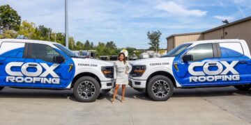 A woman in a light-colored dress and heels stands confidently between two white Ford F-150 pickup trucks with matching Cox Roofing wraps. The trucks feature bold blue and black geometric designs with the company’s name and website, coxres.com, clearly visible on the doors and hood. The scene is set in a paved parking lot in front of a commercial building under a clear blue sky.