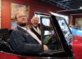 Two men are smiling while sitting inside a classic red convertible, likely a 1949 Willys Jeepster, in an indoor gallery or showroom. The younger man in the foreground wears a blue and yellow plaid blazer over a white button-down shirt and glasses. In the driver's seat, an older man with white hair wears a brown corduroy blazer and a wide grin. The background features deep red walls adorned with framed artwork and a black industrial-style ceiling with track lighting.