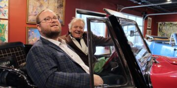 Two men are smiling while sitting inside a classic red convertible, likely a 1949 Willys Jeepster, in an indoor gallery or showroom. The younger man in the foreground wears a blue and yellow plaid blazer over a white button-down shirt and glasses. In the driver's seat, an older man with white hair wears a brown corduroy blazer and a wide grin. The background features deep red walls adorned with framed artwork and a black industrial-style ceiling with track lighting.