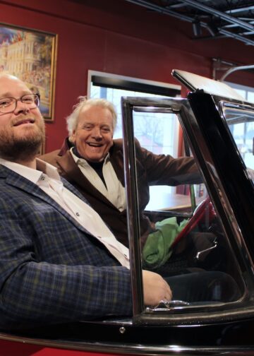 Two men are smiling while sitting inside a classic red convertible, likely a 1949 Willys Jeepster, in an indoor gallery or showroom. The younger man in the foreground wears a blue and yellow plaid blazer over a white button-down shirt and glasses. In the driver's seat, an older man with white hair wears a brown corduroy blazer and a wide grin. The background features deep red walls adorned with framed artwork and a black industrial-style ceiling with track lighting.