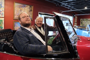 Two men are smiling while sitting inside a classic red convertible, likely a 1949 Willys Jeepster, in an indoor gallery or showroom. The younger man in the foreground wears a blue and yellow plaid blazer over a white button-down shirt and glasses. In the driver's seat, an older man with white hair wears a brown corduroy blazer and a wide grin. The background features deep red walls adorned with framed artwork and a black industrial-style ceiling with track lighting.