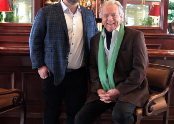 A full-length indoor photo shows two men posed together in a classic, wood-paneled bar or lounge. On the left, a younger man with glasses and a beard stands wearing a blue plaid blazer over a white button-down shirt and black trousers. On the right, an older man with silver hair sits on a leather barstool, wearing a brown blazer, a white shirt, and a long, light-green scarf draped around his neck. The background features ornate mirrors, warm lighting from wall sconces, and a large framed artwork.