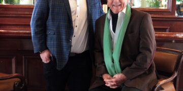 A full-length indoor photo shows two men posed together in a classic, wood-paneled bar or lounge. On the left, a younger man with glasses and a beard stands wearing a blue plaid blazer over a white button-down shirt and black trousers. On the right, an older man with silver hair sits on a leather barstool, wearing a brown blazer, a white shirt, and a long, light-green scarf draped around his neck. The background features ornate mirrors, warm lighting from wall sconces, and a large framed artwork.