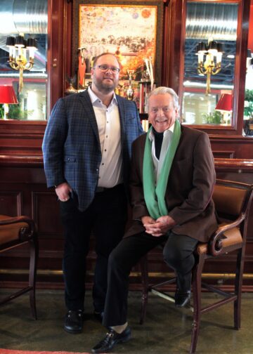 A full-length indoor photo shows two men posed together in a classic, wood-paneled bar or lounge. On the left, a younger man with glasses and a beard stands wearing a blue plaid blazer over a white button-down shirt and black trousers. On the right, an older man with silver hair sits on a leather barstool, wearing a brown blazer, a white shirt, and a long, light-green scarf draped around his neck. The background features ornate mirrors, warm lighting from wall sconces, and a large framed artwork.
