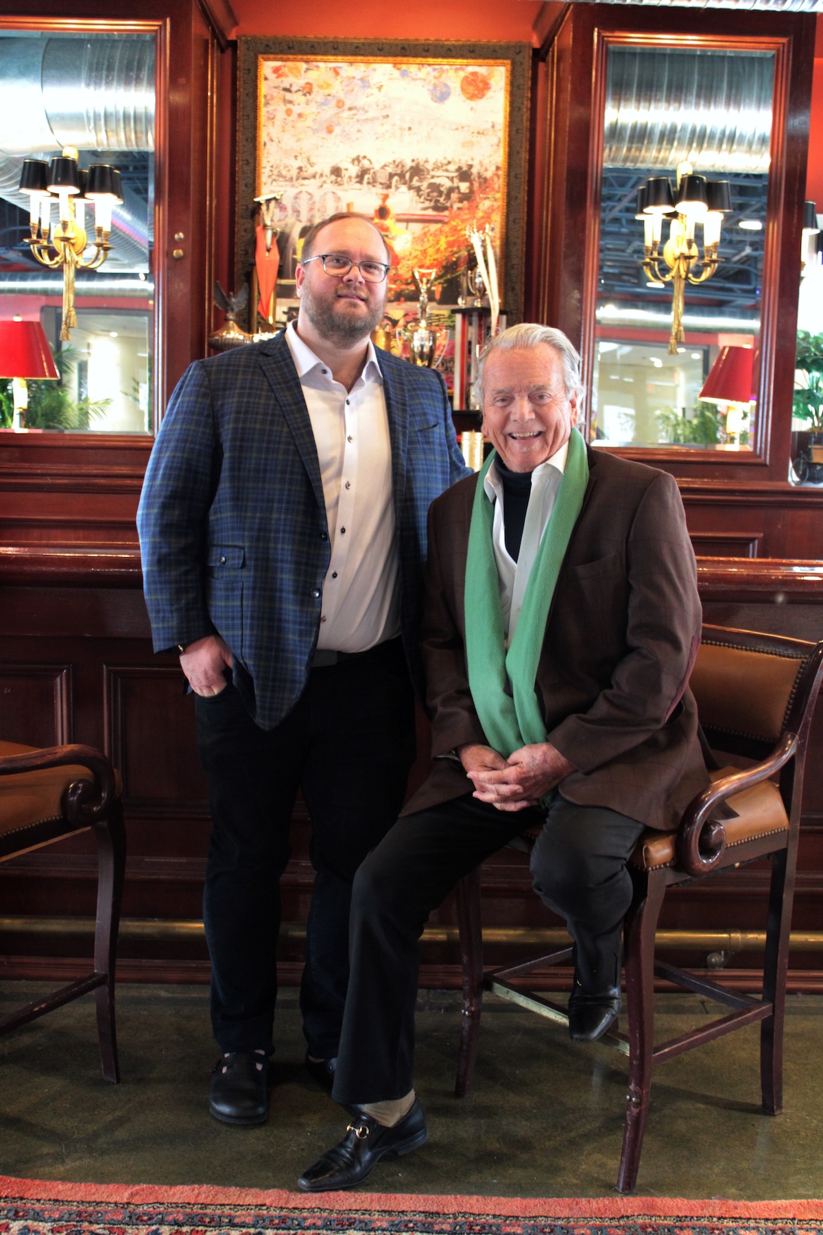 A full-length indoor photo shows two men posed together in a classic, wood-paneled bar or lounge. On the left, a younger man with glasses and a beard stands wearing a blue plaid blazer over a white button-down shirt and black trousers. On the right, an older man with silver hair sits on a leather barstool, wearing a brown blazer, a white shirt, and a long, light-green scarf draped around his neck. The background features ornate mirrors, warm lighting from wall sconces, and a large framed artwork.