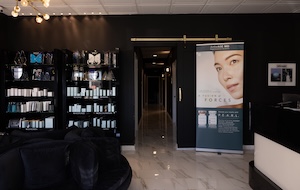 A wide shot of the modern and sophisticated lobby at an Ageless Aesthetics medical spa location. The room features dark charcoal-colored walls and polished white marble-patterned tile floors. To the left, two tall black shelving units are neatly stocked with various medical-grade skincare products. In the center, an open doorway leads down a brightly lit hallway toward treatment rooms. To the right of the doorway stands a large roll-up banner featuring a close-up of a woman's face and information about aesthetic treatments. A white reception counter is partially visible on the far right, and a circular black seating area occupies the foreground.