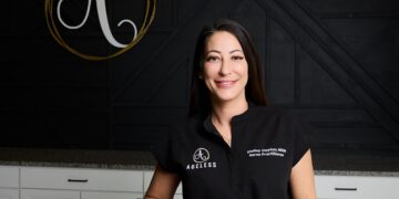 A professional studio portrait shows Shelley Clayton, MSN, FNP-BC, a board-certified nurse practitioner and founder of Ageless Aesthetics, smiling warmly at the camera. She is wearing a black collared tunic with "AGELESS" and a circular "A" logo embroidered on the right chest, and her name and title on the left. Behind her, a large gold-colored wire art piece of the same logo is mounted on a textured black wooden wall. The foreground features a sleek white counter with dark handles, suggesting a modern and high-end medical spa environment.