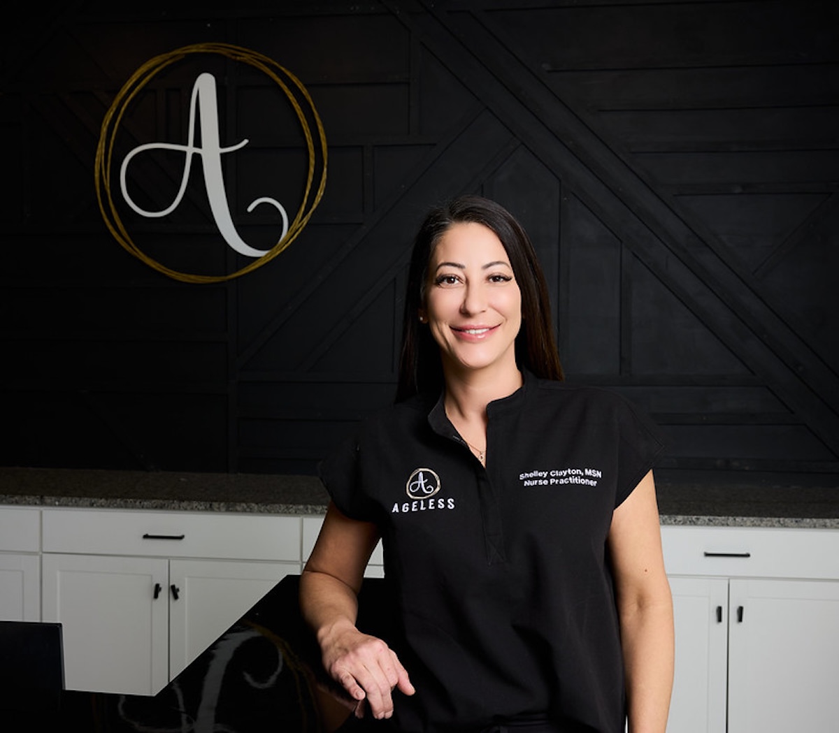 A professional studio portrait shows Shelley Clayton, MSN, FNP-BC, a board-certified nurse practitioner and founder of Ageless Aesthetics, smiling warmly at the camera. She is wearing a black collared tunic with "AGELESS" and a circular "A" logo embroidered on the right chest, and her name and title on the left. Behind her, a large gold-colored wire art piece of the same logo is mounted on a textured black wooden wall. The foreground features a sleek white counter with dark handles, suggesting a modern and high-end medical spa environment.
