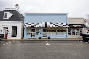 A street-level, wide shot captures a row of charming, historic storefronts on a paved street in downtown Zionsville, Indiana. On the far left is a distinctive white building with a mansard roof and a bright red door. The centerpiece is a light blue one-story building featuring a large, eye-catching black and white striped awning that spans across its front windows. The storefront on the right side of this blue building is labeled "GIFTED." To its right is a tan building with a black awning, which houses Moody's Butcher Shop. The foreground shows empty parking spaces on a damp, gray asphalt road under an overcast sky.