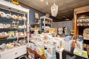 An interior view of Gifted, a boutique in Zionsville, Indiana, highlighting a thoughtfully curated children’s and baby gift section. A large white display cabinet on the left features glass shelves stocked with delicate children's books, tea sets, and decorative items. In the center, a white table is covered with an array of gift-boxed toys and plush animals. To the right, a wooden shelf unit displays additional treasures, while a blue curtained area in the background reveals a selection of infant and children's clothing. The shop features a modern-vintage aesthetic with dark blue walls, a white patterned tin ceiling, and a large, woven pendant light hanging from the center.