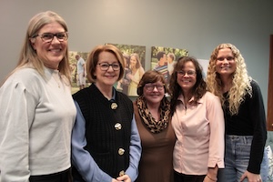 A diverse group of five smiling women stand together for a group photo in a modern, brightly lit office or gallery space. The women are arranged in a single row, with most wearing casual professional attire, including a light blue sweater, a black knit vest, a brown top with a leopard-print scarf, a light pink blouse, and a black long-sleeved top with jeans. In the background, four large canvas prints of family and couple portraits are mounted on a gray wall.