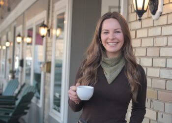 A medium shot of a smiling woman with long brown hair, wearing a dark brown long-sleeved top and an olive green polka-dot bandana tied around her neck. She is holding a white ceramic coffee cup in her right hand and is standing outdoors in front of a light-colored brick wall. To her left, a row of lit, classic-style black lanterns is mounted on the side of a building, with several dark green Adirondack chairs visible in the softly blurred background along a porch.