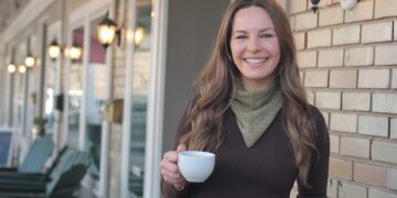 A medium shot of a smiling woman with long brown hair, wearing a dark brown long-sleeved top and an olive green polka-dot bandana tied around her neck. She is holding a white ceramic coffee cup in her right hand and is standing outdoors in front of a light-colored brick wall. To her left, a row of lit, classic-style black lanterns is mounted on the side of a building, with several dark green Adirondack chairs visible in the softly blurred background along a porch.