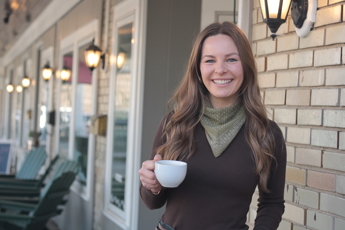 A medium shot of a smiling woman with long brown hair, wearing a dark brown long-sleeved top and an olive green polka-dot bandana tied around her neck. She is holding a white ceramic coffee cup in her right hand and is standing outdoors in front of a light-colored brick wall. To her left, a row of lit, classic-style black lanterns is mounted on the side of a building, with several dark green Adirondack chairs visible in the softly blurred background along a porch.
