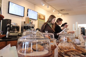 A bustling scene inside Roasted in the Village in Zionsville, Indiana. Two women with long dark hair, dressed in black, are working behind a dark wooden counter. One appears to be using a tablet at the register, while the other is focused on a task nearby. In the foreground, several large glass cake domes on the counter display fresh pastries and cookies. A basket of breadsticks is also visible to the right. The background features a clean, white-walled interior with two large digital menu screens and professional coffee equipment, including an espresso machine and grinder, all under bright, warm lighting.