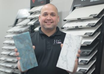 A medium shot of a smiling man with a shaved head and a short beard, wearing a black polo shirt with a white circular logo that reads "JC COUNTERTOPS - MARBLE • GRANITE • QUARTZ." He is holding up two rectangular stone samples: a vibrant, swirled blue and green stone in his right hand and a white stone with delicate gray veining (resembling Carrara marble) in his left. He stands in a showroom in front of several black display racks filled with various labeled stone and tile samples, including "MSI" and "Surfaces."