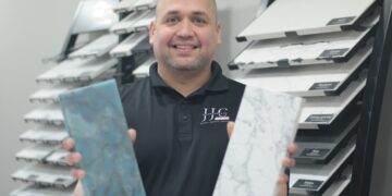 A medium shot of a smiling man with a shaved head and a short beard, wearing a black polo shirt with a white circular logo that reads "JC COUNTERTOPS - MARBLE • GRANITE • QUARTZ." He is holding up two rectangular stone samples: a vibrant, swirled blue and green stone in his right hand and a white stone with delicate gray veining (resembling Carrara marble) in his left. He stands in a showroom in front of several black display racks filled with various labeled stone and tile samples, including "MSI" and "Surfaces."