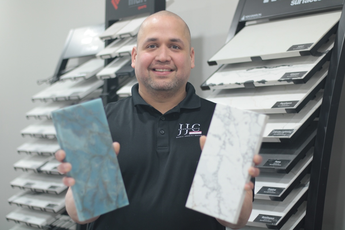 A medium shot of a smiling man with a shaved head and a short beard, wearing a black polo shirt with a white circular logo that reads "JC COUNTERTOPS - MARBLE • GRANITE • QUARTZ." He is holding up two rectangular stone samples: a vibrant, swirled blue and green stone in his right hand and a white stone with delicate gray veining (resembling Carrara marble) in his left. He stands in a showroom in front of several black display racks filled with various labeled stone and tile samples, including "MSI" and "Surfaces."