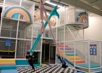 A bright and whimsical indoor playground designed with a soft-colored rainbow theme. The centerpiece is a multi-level white mesh climbing structure that includes padded platforms, a beige tube tunnel, and large circular viewing portals decorated with rainbow graphics. In the foreground, two children are actively swinging on long, pink and teal aerial silk fabrics over a black-and-white checkered padded mat. To the right, a set of padded stairs in a spectrum of pastel colors leads up into the play structure. The facility has an open, industrial-style ceiling and is safety-oriented with white mesh netting throughout.