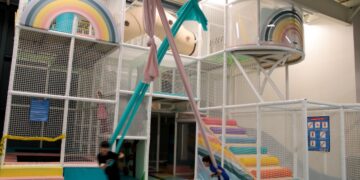 A bright and whimsical indoor playground designed with a soft-colored rainbow theme. The centerpiece is a multi-level white mesh climbing structure that includes padded platforms, a beige tube tunnel, and large circular viewing portals decorated with rainbow graphics. In the foreground, two children are actively swinging on long, pink and teal aerial silk fabrics over a black-and-white checkered padded mat. To the right, a set of padded stairs in a spectrum of pastel colors leads up into the play structure. The facility has an open, industrial-style ceiling and is safety-oriented with white mesh netting throughout.