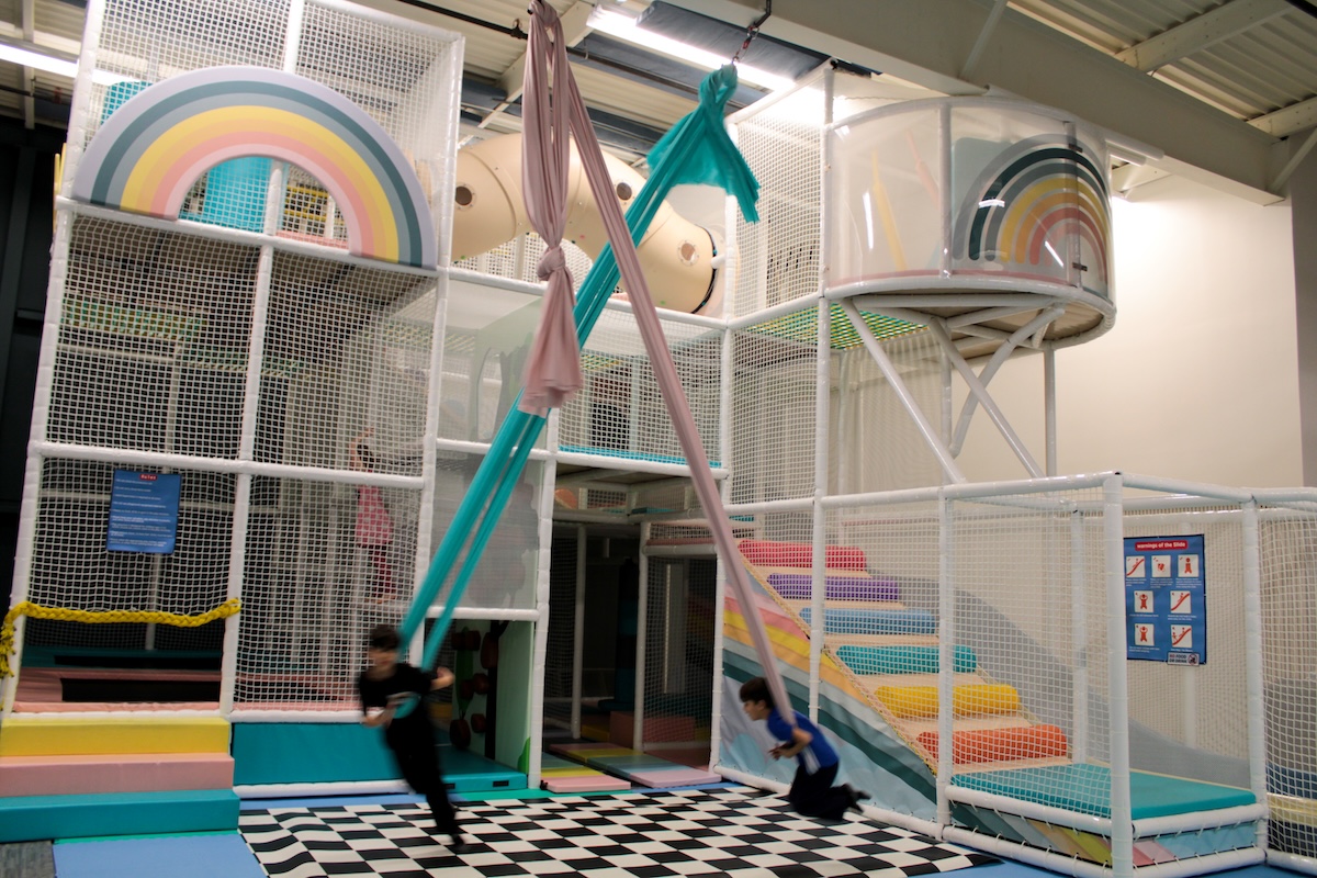 A bright and whimsical indoor playground designed with a soft-colored rainbow theme. The centerpiece is a multi-level white mesh climbing structure that includes padded platforms, a beige tube tunnel, and large circular viewing portals decorated with rainbow graphics. In the foreground, two children are actively swinging on long, pink and teal aerial silk fabrics over a black-and-white checkered padded mat. To the right, a set of padded stairs in a spectrum of pastel colors leads up into the play structure. The facility has an open, industrial-style ceiling and is safety-oriented with white mesh netting throughout.