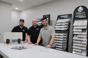 Three smiling men stand behind a polished white stone island in a modern countertop showroom. The two men on the left wear black polo shirts with the "JC COUNTERTOPS" logo, which also lists "MARBLE • GRANITE • QUARTZ." The man on the right wears a light gray polo shirt. To the left, a stone sample is displayed on a stand. The background features several large, black display racks filled with various stone and tile samples, including prominent branding for "mont SURFACES" and "FROM MSI." The room is brightly lit with recessed ceiling lights against neutral gray walls.