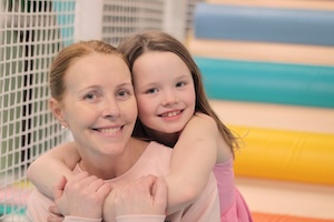 A close-up, heartwarming portrait shows a smiling woman being hugged from behind by a young girl. The woman, with her light brown hair pulled back, is wearing a light pink long-sleeved top. The girl, also smiling broadly, has long brown hair and is wearing a pink dress. They are in a brightly lit indoor play area, with a white mesh safety net to the left and a wall of colorful, horizontal padded tubes in shades of blue, teal, and yellow in the soft-focus background.