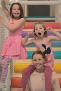 A joyful portrait of a woman and two young girls posing on a set of colorful, padded indoor playground stairs. The woman, with her red-brown hair pulled back, is smiling warmly at the camera while wearing a light pink sweatshirt. One girl, in a pink sleeveless dress and lavender leggings, stands to her left and leans toward her. The other girl, wearing a pink Mickey Mouse tank top and polka-dot leggings, is perched playfully on the woman's shoulders, making a funny face with her mouth open and fingers in her ears. The background features a vibrant spectrum of large, horizontal padded tubes in shades of orange, yellow, teal, blue, and pink.