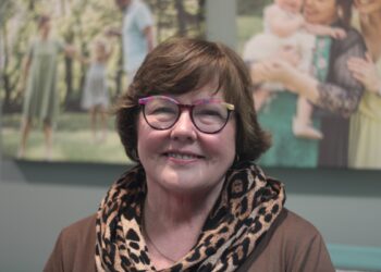 A close-up portrait of a woman with short, wavy auburn hair and glasses with colorful, patterned frames. She is smiling warmly at the camera and wearing a brown top paired with a voluminous leopard-print infinity scarf. In the softly blurred background, two large canvas prints of family photographs are mounted on a gray wall. One print on the left shows a group in a park-like setting, and the one on the right features a woman holding a baby.