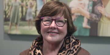 A close-up portrait of a woman with short, wavy auburn hair and glasses with colorful, patterned frames. She is smiling warmly at the camera and wearing a brown top paired with a voluminous leopard-print infinity scarf. In the softly blurred background, two large canvas prints of family photographs are mounted on a gray wall. One print on the left shows a group in a park-like setting, and the one on the right features a woman holding a baby.