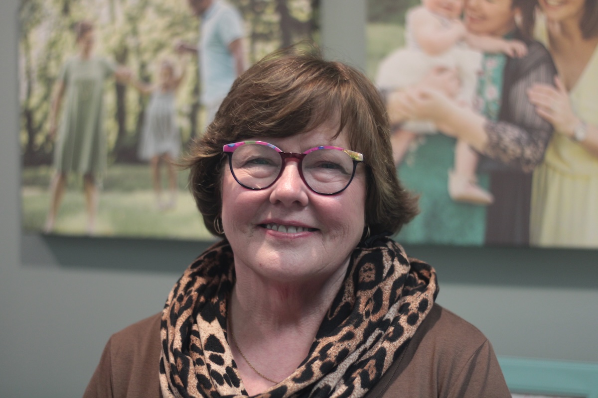 A close-up portrait of a woman with short, wavy auburn hair and glasses with colorful, patterned frames. She is smiling warmly at the camera and wearing a brown top paired with a voluminous leopard-print infinity scarf. In the softly blurred background, two large canvas prints of family photographs are mounted on a gray wall. One print on the left shows a group in a park-like setting, and the one on the right features a woman holding a baby.