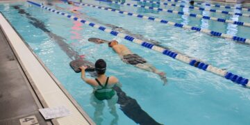 An indoor swimming pool with several lap lanes marked by blue and white buoy lines. In the foreground, a woman in a green swimsuit and a man in black trunks are performing training drills using black kickboards. The pool deck features a depth marker for "4 FT 4 IN" and a "NO DIVING" symbol, with a printed workout sheet resting nearby. The background shows a bright, modern facility with an orange rescue board mounted on the wall, a white lifeguard stand, and other swimmers in the distance.