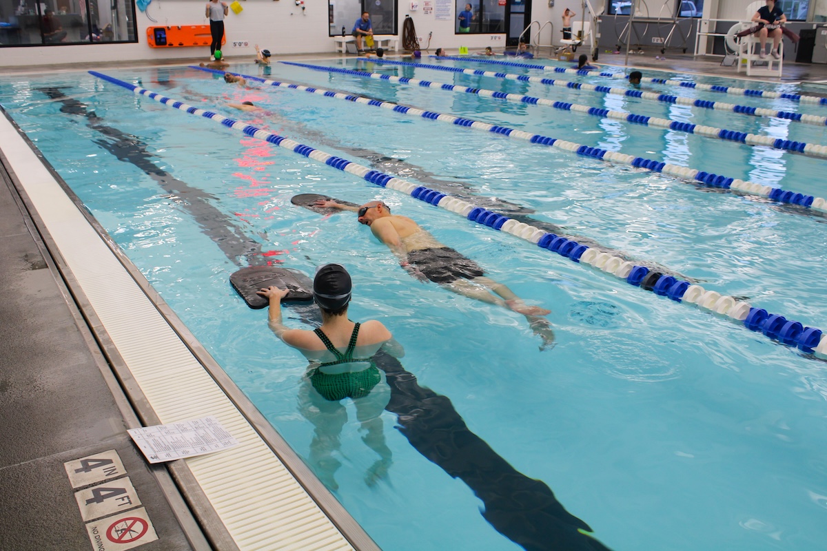An indoor swimming pool with several lap lanes marked by blue and white buoy lines. In the foreground, a woman in a green swimsuit and a man in black trunks are performing training drills using black kickboards. The pool deck features a depth marker for "4 FT 4 IN" and a "NO DIVING" symbol, with a printed workout sheet resting nearby. The background shows a bright, modern facility with an orange rescue board mounted on the wall, a white lifeguard stand, and other swimmers in the distance.