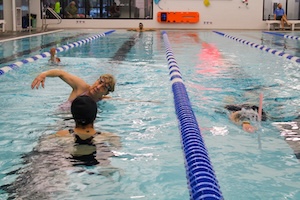 An action shot in an indoor swimming pool shows several swimmers in various lanes during a practice or training session. In the center lane, a young male swimmer wearing goggles is captured mid-stroke, his right arm reaching forward and creating a splash. In the foreground, the back of a woman in a black swimsuit and swim cap is visible as she watches or coaches from the lane. In the lane to the right, a swimmer is partially submerged, while another swimmer is visible further down the pool. Blue and white lane lines divide the pool, and an orange rescue board is mounted on the far wall of the brightly lit facility.