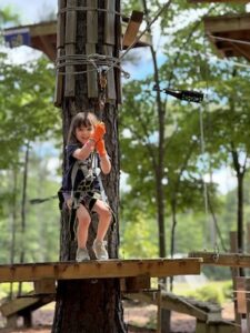 A medium shot of a young girl with dark hair and a wide smile, wearing orange safety gloves and a full-body climbing harness. She is suspended on a zipline, holding onto a black rope as she begins her ride from a wooden platform attached to a tall pine tree. The background is a lush, out-of-focus green forest under a bright, sunny sky. The zipline and safety cables are visible above her, and other wooden platforms are seen in the distance, part of a high-ropes adventure course.
